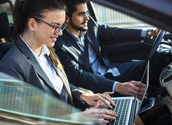 Two people in the front seat of a car. The man is driving, the woman is working on her laptop.