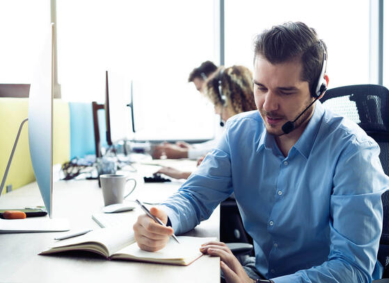Man with headset writing in a book while working at a contact center