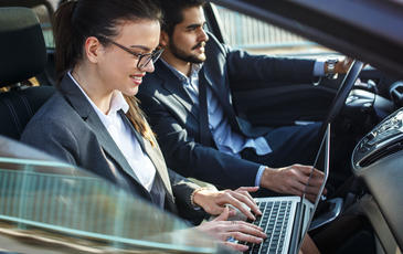 Two people in the front seat of a car. The man is driving, the woman is working on her laptop.