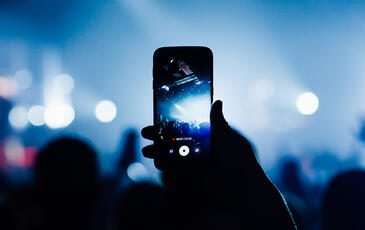 Close-up of a mobile phone recording a live concert, with blurred performers and audience visible on the phone screen