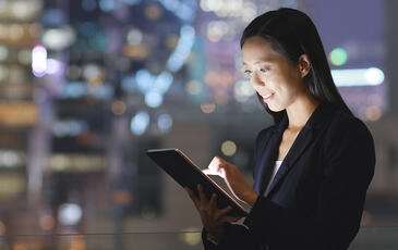 Asian businesswoman in a bustling city at night, using a tablet with city lights glowing around her
