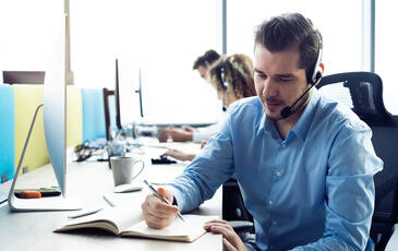Man with headset writing in a book while working at a contact center