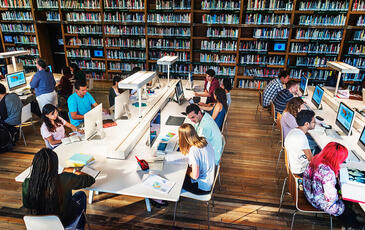 Several students working in a technology library, using computers, books, and printed materials.