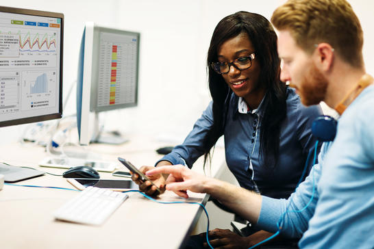 Man and woman discussing while looking at mobile phone and computer screens