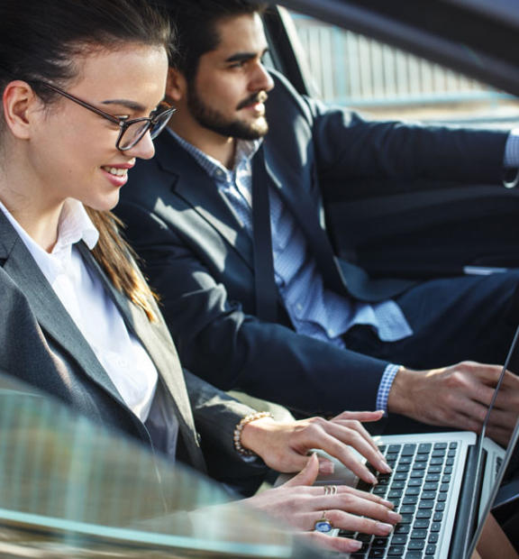 Two people in the front seat of a car. The man is driving, the woman is working on her laptop.
