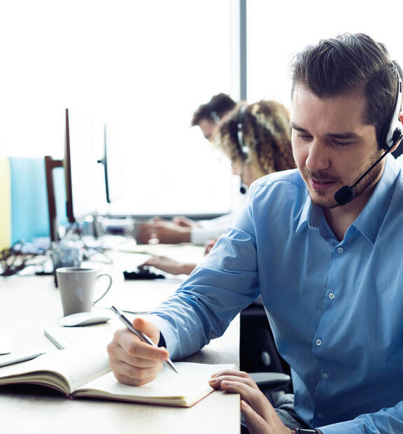 Man with headset writing in a book while working at a contact center