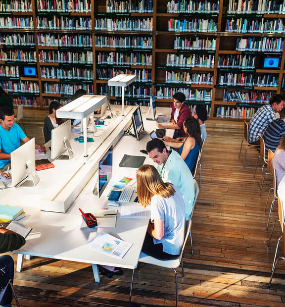 Several students working in a technology library, using computers, books, and printed materials.