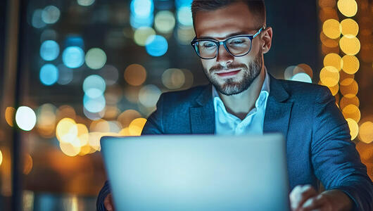 Young professional working late on laptop with city night lights, symbolizing Non-Terrestrial Networks (NTNs) innovation.